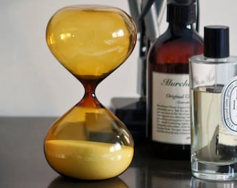 May include: A yellow hourglass with a bulbous top and bottom. The hourglass is filled with sand and is sitting on a black surface.