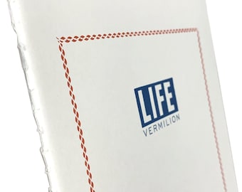 Op de afbeelding: Een wit notitieboek met een handgestikte rug en een rode rand met een touwpatroon. De kaft heeft een blauw rechthoekig logo met het woord LIFE in dikke hoofdletters, met daaronder het woord VERMILION in kleinere letters.
