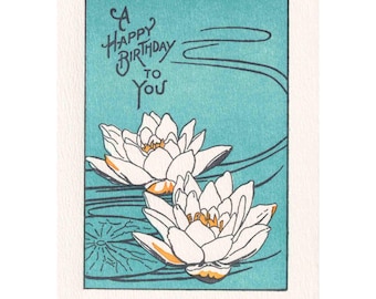 May include: A greeting card with a teal background and white border. The card features two white water lilies with yellow centers. The text "A Happy Birthday to You" is written in a script font above the flowers.