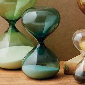 May include: Two green glass hourglasses with white sand. The hourglass on the left is larger than the hourglass on the right.