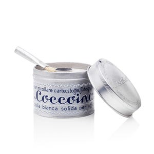 May include: A round silver metal tin of Coccoina white solid glue with a matching lid placed to the side. The label features blue text reading per incollare carte, stoffe, fotografie and Coccoina colla bianca solida per ufficio. A small brush is inside.