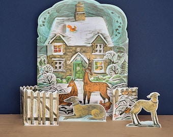 May include: A die-cut cardboard advent calendar featuring a snow-covered cottage with a green door, surrounded by a deer, a squirrel, and a chicken. In the foreground, a separate cutout shows a tan dog lying down and another standing dog.
