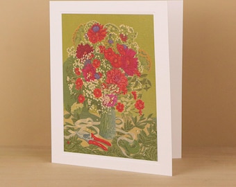 May include: A white greeting card featuring a printed illustration of a vibrant bouquet of red and pink flowers in a glass vase, set against a muted olive green background. Red garden shears and white ribbon are placed on the surface below.