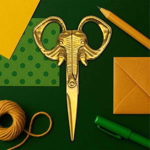 Puede incluir: Tijeras doradas con diseño de cabeza de elefante, sobre un fondo verde con artículos de papelería. Las tijeras son el punto focal, con una cara de elefante detallada que forma las asas. Otros artículos incluyen un lápiz, un bolígrafo y papel.