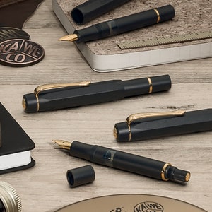Könnte beinhalten: Vier schwarze Füllfederhalter mit goldenen Akzenten und Federn. Die Stifte liegen auf einer Holzoberfläche mit einem Notizbuch und anderen Gegenständen.
