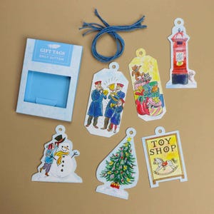 May include: A set of Emily Sutton gift tags featuring a light blue box, a coil of blue twine, and six illustrated tags depicting a snowman, a Christmas tree, a toy shop sign, a red post box, a group of musicians, and a stack of gift boxes.
