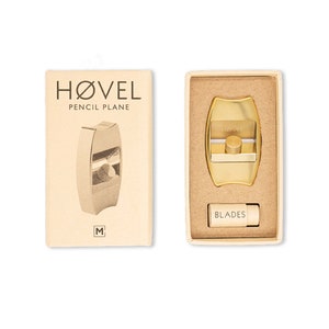 Op de afbeelding: Een gouden metalen potloodpuntenslijper met het woord "HOVEL" erop. De puntenslijper zit in een kartonnen doos met de woorden "PENCIL PLANE" en "HOVEL" erop. De doos bevat ook een klein doosje met vervangende messen met het label "BLADES".