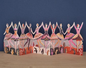 May include: A decorative paper accordion card featuring a continuous line of ten illustrated dancers holding hands. Each figure wears a unique, colorful ballet-style outfit with intricate patterns, set against a stylized landscape of foliage.