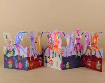 May include: A multi-paneled accordion-fold card featuring a vibrant, colorful illustration of anthropomorphic animals and figures in elaborate costumes dancing together. The scene includes foxes, birds, and masked characters amidst stylized foliage.
