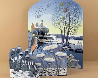 May include: A multi-layered paper advent calendar featuring a winter landscape with a house, a kingfisher bird, snowdrops, and a swan. The scene includes numbered circles for counting down days, set against a blue and yellow sky with bare trees.