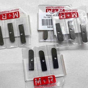 May include: Three clear plastic wallets each containing three dark grey metal replacement blades for pencil sharpeners. Each blade features a single circular hole and is branded with the M+R Möbiu+Ruppert logo in red and white text on the packaging.