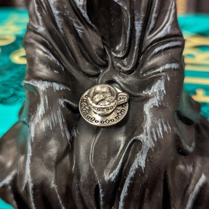 May include: A silver teacup and saucer charm with intricate details. The charm is attached to a black fabric background.