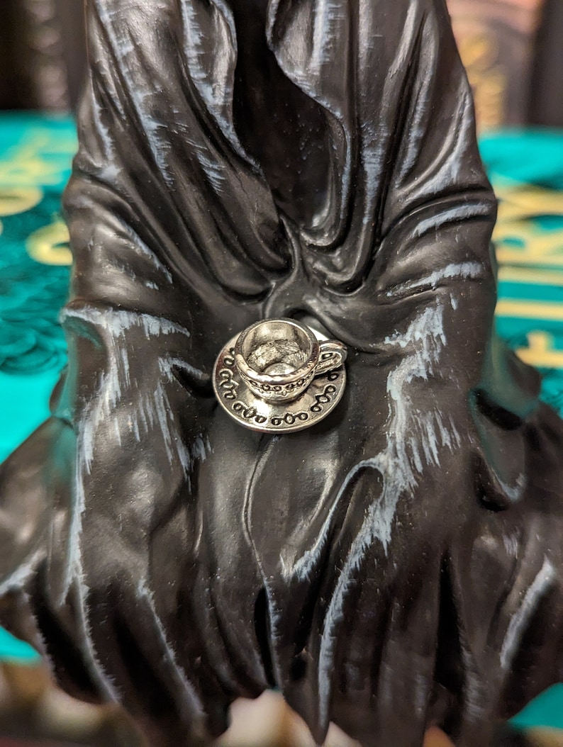 May include: A silver teacup and saucer charm with intricate details. The charm is attached to a black fabric background.