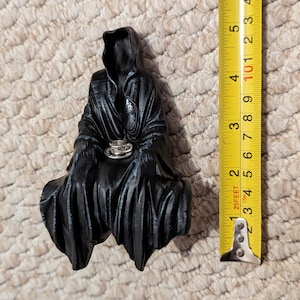 May include: Black figurine of a hooded figure sitting with a small teacup in their hands. The figurine is approximately 5 inches tall.