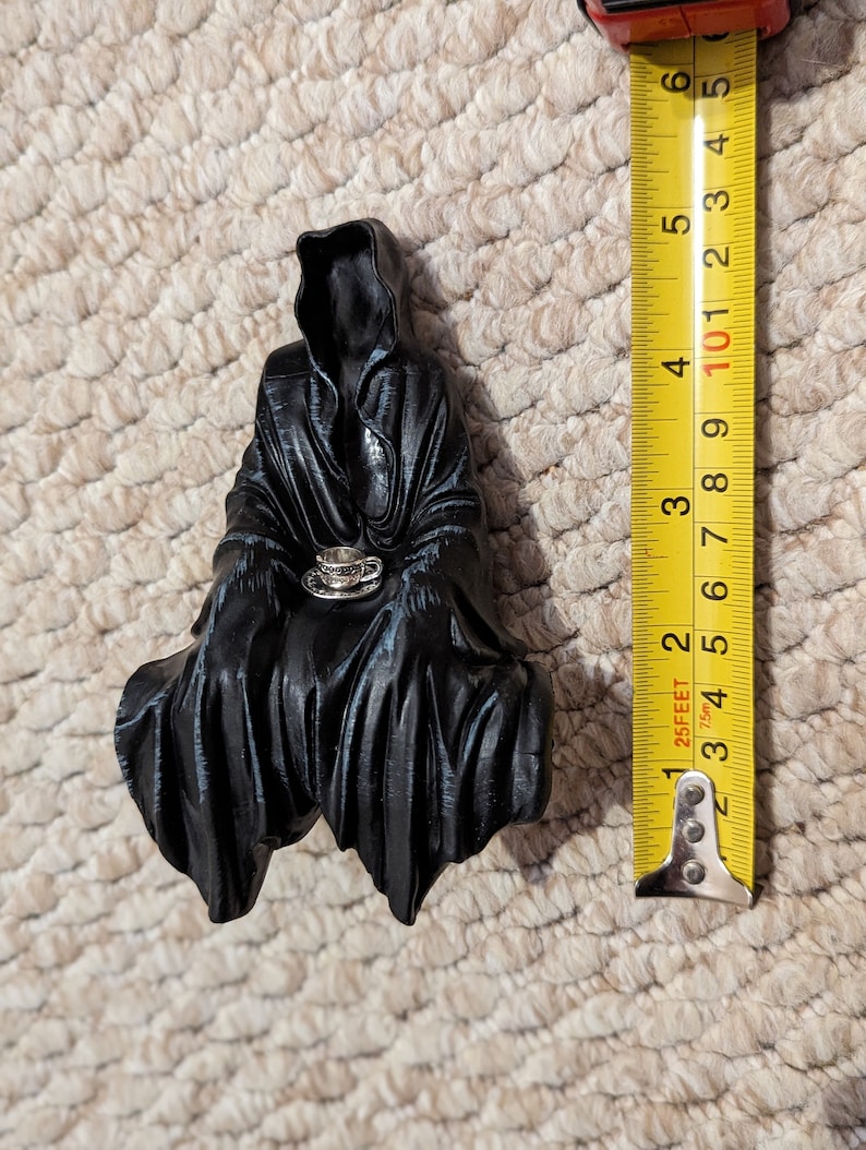 May include: Black figurine of a hooded figure sitting with a small teacup in their hands. The figurine is approximately 5 inches tall.