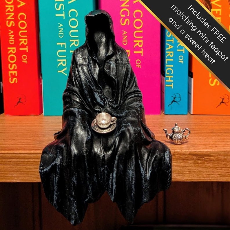 May include: A black figurine of a hooded figure sitting with a teacup in its lap. The figurine is on a shelf with a row of colorful books behind it. A small silver teapot is on the shelf to the right of the figurine. The text "Includes FREE matching mini teapot and a sweet treat" is in a white box with a black border.