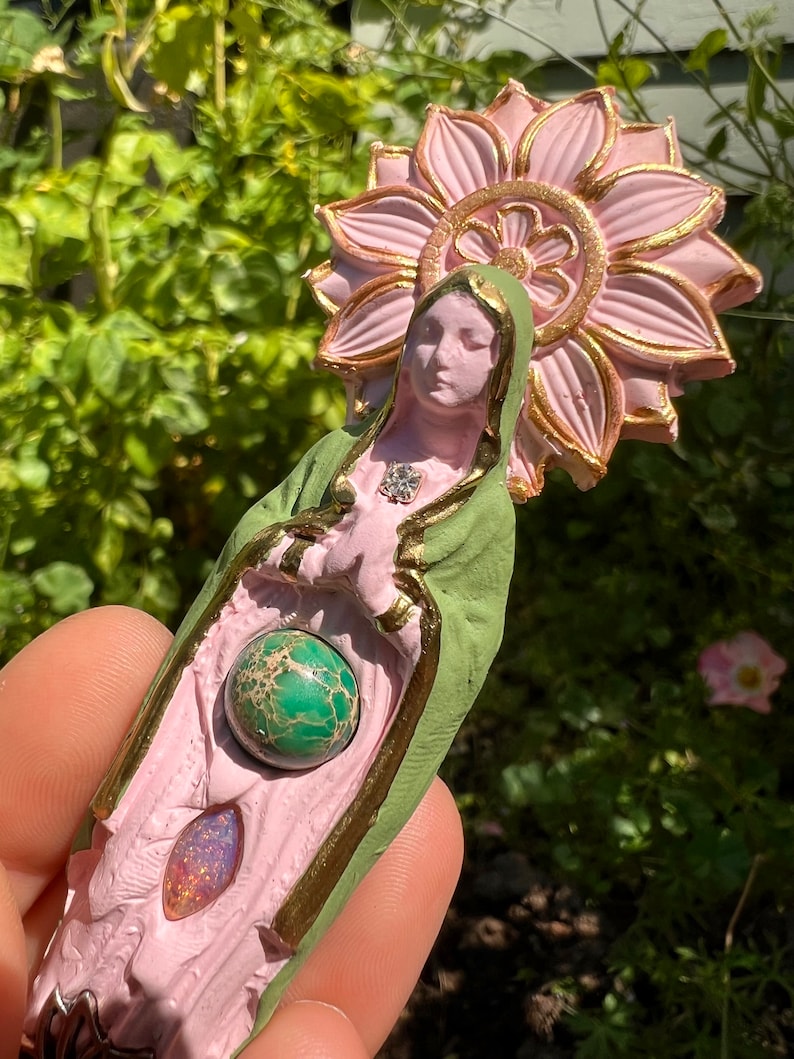 May include: A pink and green figurine of a woman with a flower halo. The figurine has a green gemstone in its hands and a pink gemstone in its lap.