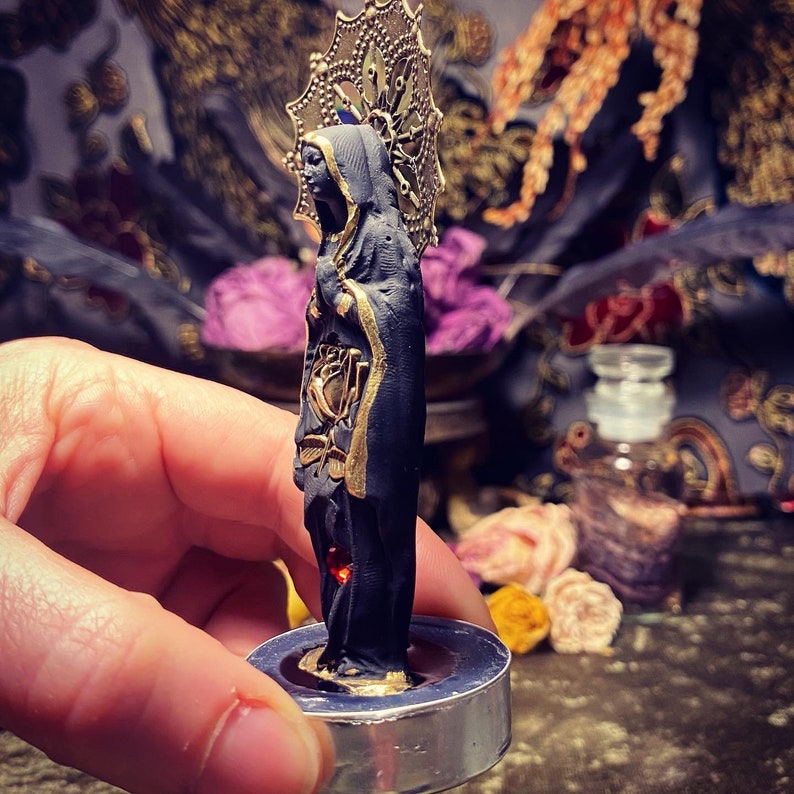 May include: A black and gold figurine of a woman wearing a robe and a crown. The figurine is standing on a round, silver base.