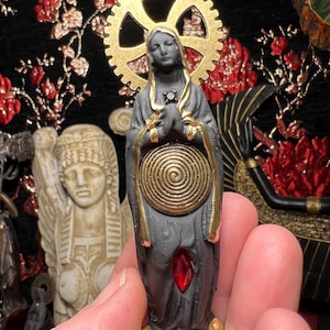 May include: A small, dark gray statue of a figure with a gold halo and accents. The statue features a spiral design on the chest and a red gem. The base is gold. The figure is holding its hands together in a prayer position.