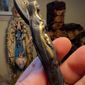 Puede incluir: Peque&ntilde;a estatua negra de una figura vestida con una t&uacute;nica, con un halo y detalles dorados. La figura sostiene un objeto circular con un dise&ntilde;o dorado. La estatua tiene una expresi&oacute;n serena y es probablemente un art&iacute;culo religioso o espiritual.