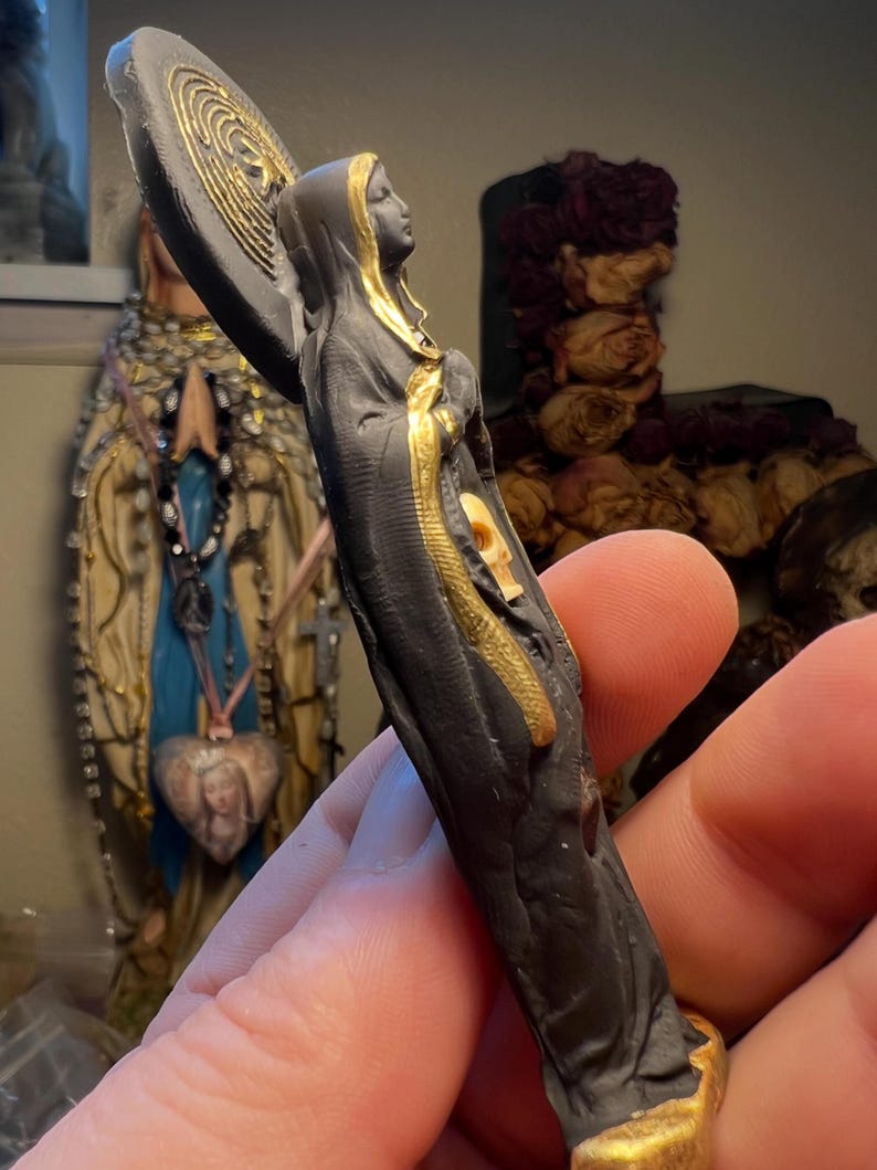 Puede incluir: Peque&ntilde;a estatua negra de una figura vestida con una t&uacute;nica, con un halo y detalles dorados. La figura sostiene un objeto circular con un dise&ntilde;o dorado. La estatua tiene una expresi&oacute;n serena y es probablemente un art&iacute;culo religioso o espiritual.