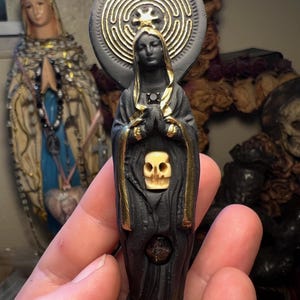 May include: A black and gold statue of a figure with a skull on the chest and a labyrinth design behind the head. The statue is held in a hand, with another statue in the background.