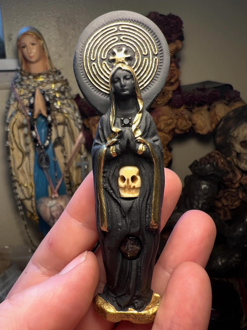 Puede incluir: Una estatua negra y dorada de una figura con una calavera en el pecho y un dise&ntilde;o de laberinto detr&aacute;s de la cabeza. La estatua se sostiene en una mano, con otra estatua en el fondo.