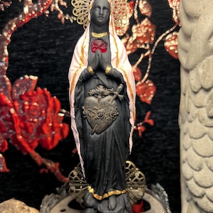 Op de afbeelding: Zwarte figuur van de Maagd Maria met een gouden halo en een rood hart op haar borst. Het beeldje staat op een zwarte basis met gouden accenten.