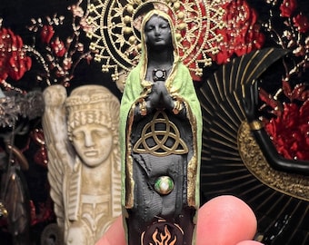 Puede incluir: Una pequeña estatua de una figura con las manos juntas en oración, vestida con un chal verde y una túnica negra. La estatua tiene un halo dorado y adornos dorados. Un símbolo dorado y un símbolo de llama están en la parte delantera de la túnica. La estatua se sostiene en la mano de una persona.