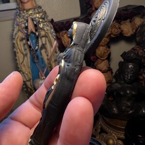 Puede incluir: Peque&ntilde;a figura religiosa de color oscuro con detalles dorados. La figura lleva una t&uacute;nica con capucha y tiene un dise&ntilde;o de halo detr&aacute;s de la cabeza. La figura se sostiene en una mano, con otros objetos religiosos en el fondo.