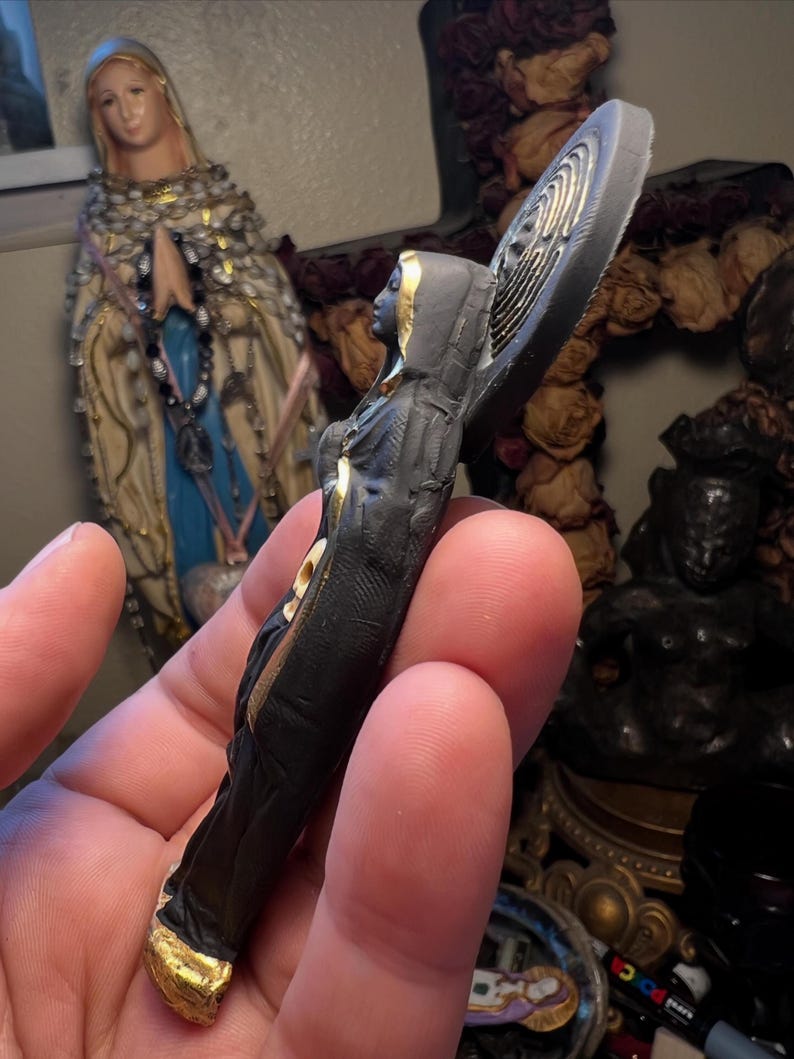 Puede incluir: Peque&ntilde;a figura religiosa de color oscuro con detalles dorados. La figura lleva una t&uacute;nica con capucha y tiene un dise&ntilde;o de halo detr&aacute;s de la cabeza. La figura se sostiene en una mano, con otros objetos religiosos en el fondo.
