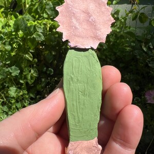 May include: A hand-painted ceramic figurine of a person with a pink flower head and a green dress.