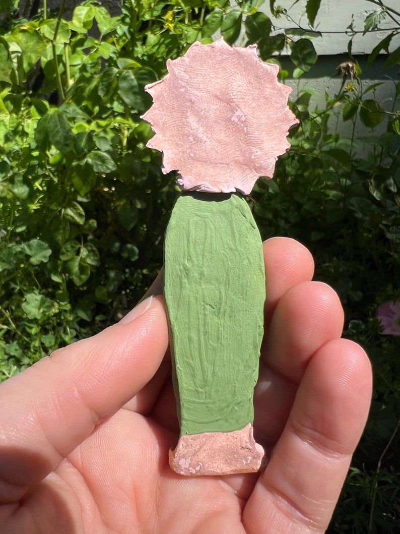 May include: A hand-painted ceramic figurine of a person with a pink flower head and a green dress.