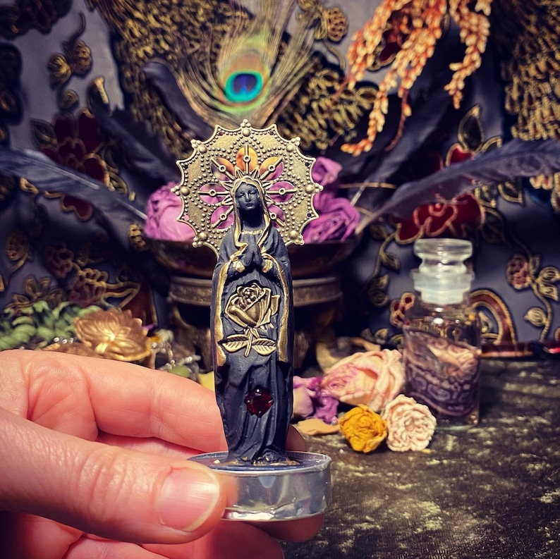 May include: Black figurine of a woman with a gold halo and a rose in her hand. The figurine is standing on a clear glass base.