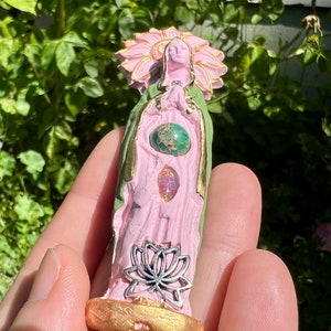 May include: A pink and green ceramic figurine of a woman with a flower crown, wearing a green robe and holding a silver lotus flower. The figurine has three gemstones embedded in the chest area.