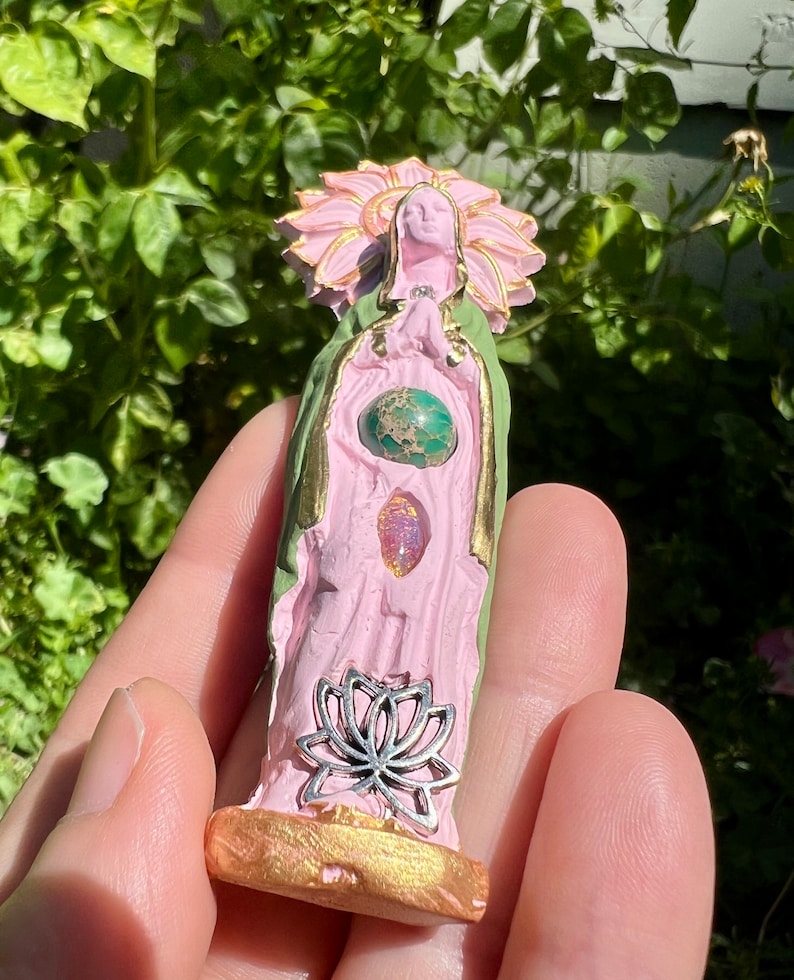May include: A pink and green ceramic figurine of a woman with a flower crown, wearing a green robe and holding a silver lotus flower. The figurine has three gemstones embedded in the chest area.