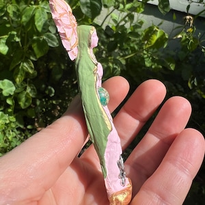 May include: A green and pink figurine of a woman with a flower crown and a green gemstone in her belly. The figurine is made of paper mache and has a gold finish.