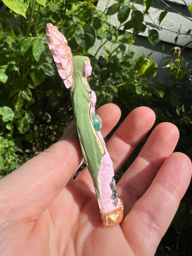 May include: A green and pink figurine of a woman with a flower crown and a green gemstone in her belly. The figurine is made of paper mache and has a gold finish.