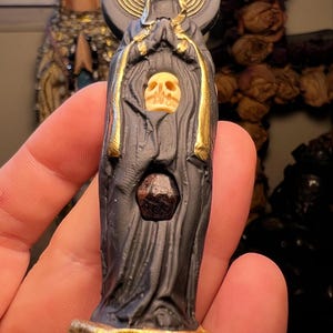 Puede incluir: Estatua oscura de una figura vestida con una t&uacute;nica con una calavera y una piedra preciosa. La estatua tiene detalles dorados y un halo con un dise&ntilde;o de espiral dorada. Las manos de la figura est&aacute;n juntas en oraci&oacute;n.