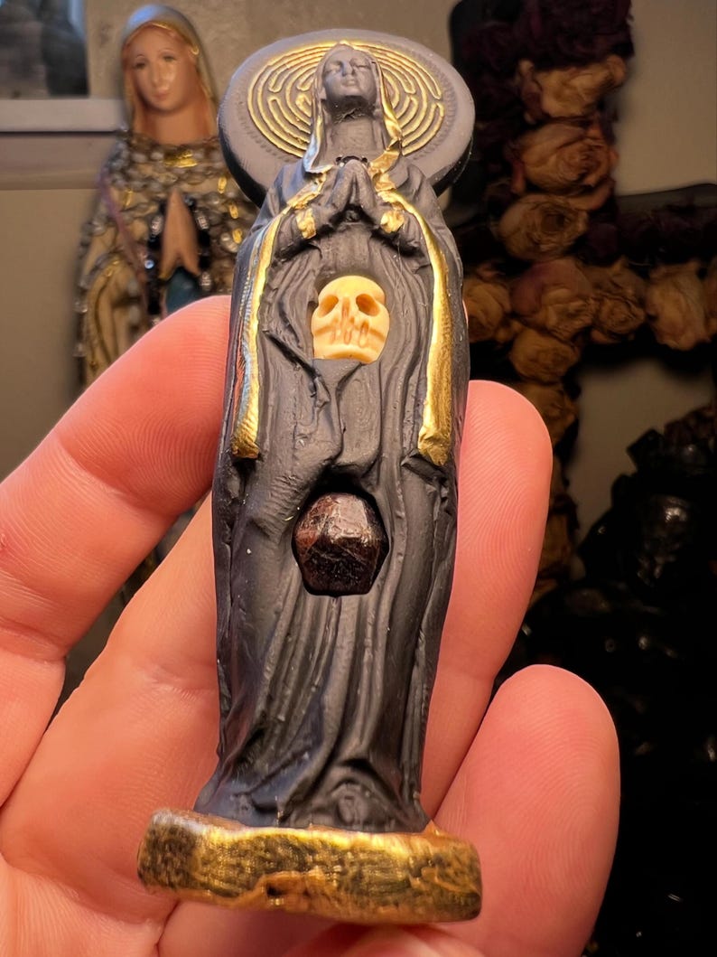 Puede incluir: Estatua oscura de una figura vestida con una t&uacute;nica con una calavera y una piedra preciosa. La estatua tiene detalles dorados y un halo con un dise&ntilde;o de espiral dorada. Las manos de la figura est&aacute;n juntas en oraci&oacute;n.