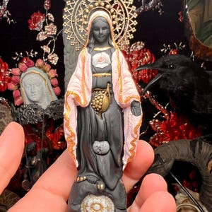 Puede incluir: Figurilla de cerámica negra y rosa de la Virgen María con un halo dorado y un corazón con un ojo en el pecho. La figurilla está de pie sobre una base con una flor blanca.