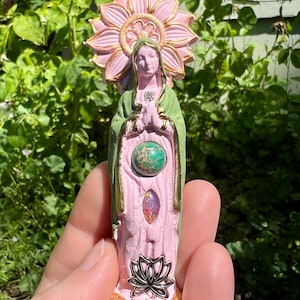 May include: A pink and green ceramic statue of a woman with a halo of pink flowers. The statue has a green robe and a pink dress. The woman is holding her hands together in prayer. The statue has three gemstones embedded in the front.