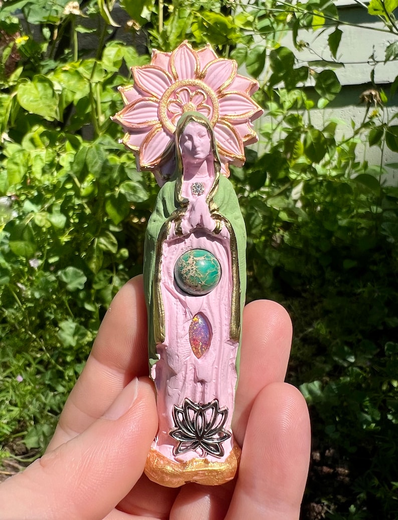 May include: A pink and green ceramic statue of a woman with a halo of pink flowers. The statue has a green robe and a pink dress. The woman is holding her hands together in prayer. The statue has three gemstones embedded in the front.