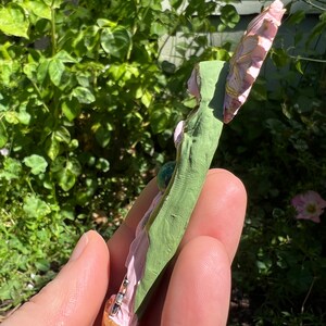 May include: A pink and green fairy figurine with a gold base and a green gemstone. The fairy is holding a flower in her hand.