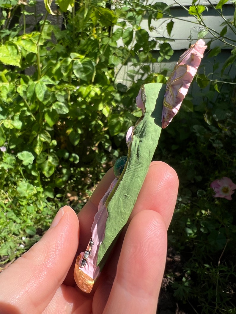 May include: A pink and green fairy figurine with a gold base and a green gemstone. The fairy is holding a flower in her hand.