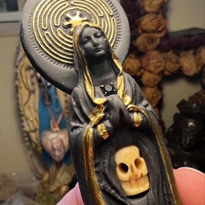 Puede incluir: Estatua negra de una figura vestida con detalles dorados, una calavera y un halo circular con un dise&ntilde;o de laberinto dorado. Las manos de la figura est&aacute;n juntas en oraci&oacute;n. Un art&iacute;culo religioso o espiritual.