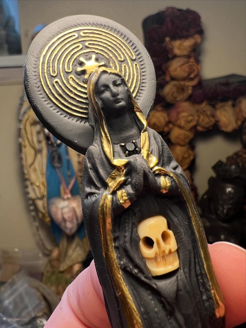 Puede incluir: Estatua negra de una figura vestida con detalles dorados, una calavera y un halo circular con un dise&ntilde;o de laberinto dorado. Las manos de la figura est&aacute;n juntas en oraci&oacute;n. Un art&iacute;culo religioso o espiritual.