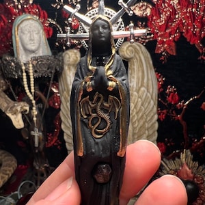 May include: Black figurine of a woman wearing a black robe with gold trim and a gold dragon on her chest. She is wearing a crown of swords and has a black face. The figurine is standing on a gold base.