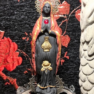 Op de afbeelding: Zwarte figuur van een vrouw die een rode en gouden jurk draagt met een kroon van gouden bladeren. De figuur houdt een gouden kelk en een rode edelsteen vast. De figuur staat op een zwarte basis met een schedel.