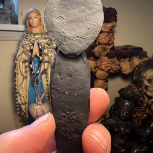 Puede incluir: Una figura hecha a mano, de color gris oscuro, con una cabeza grande y redonda y una superficie texturizada. La figura tiene un cuerpo largo y rectangular y una peque&ntilde;a base. La figura se sostiene en la mano de una persona, con otros objetos religiosos en el fondo.